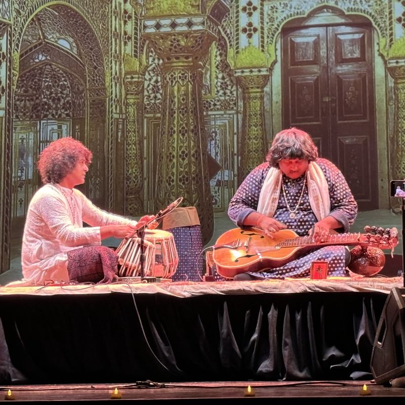 Cassius Khan and Salil Bhatt perform on stage with a projection of an ornately decorated Indian hall in the background. 