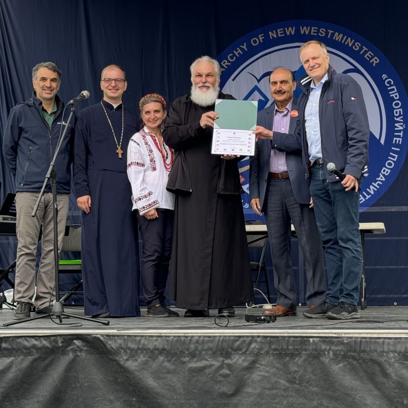 Patrick, Peter Julian and Raj Chouhan stand on a stage with representatives of the Ukrainian Catholic Eparchy of New Westminster.