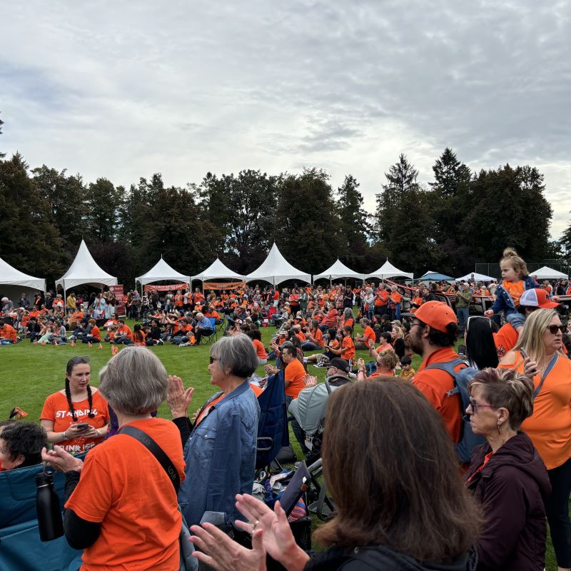 Hundreds of people, most in orange shirts, assemble on a grass field in Queens Park. 