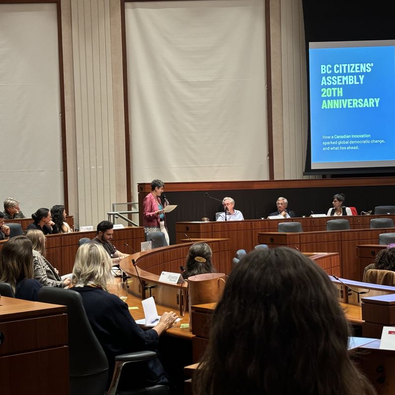 People meet in the round auditorium of the Wosk Centre for Dialogue as a woman speaks into a microphone and a screen behind reads "BC Citizens' Assembly 20th Anniversary" 