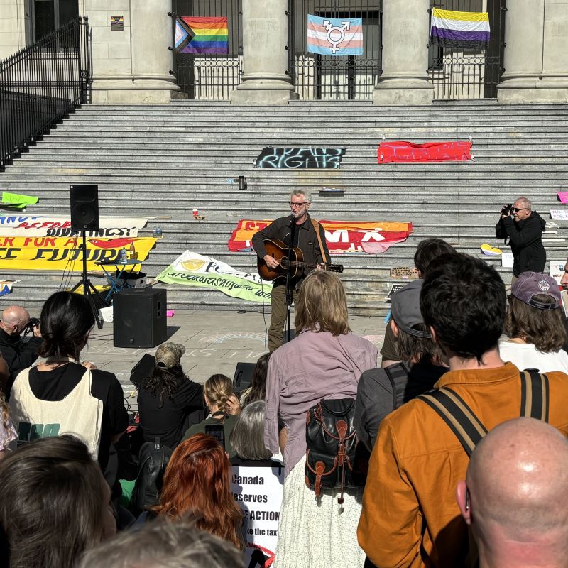 Billy Brag plays guitar and sings, alone at a microphone in front of the Vancouver Art Gallery steps with protest flags around and a crowd watching. 