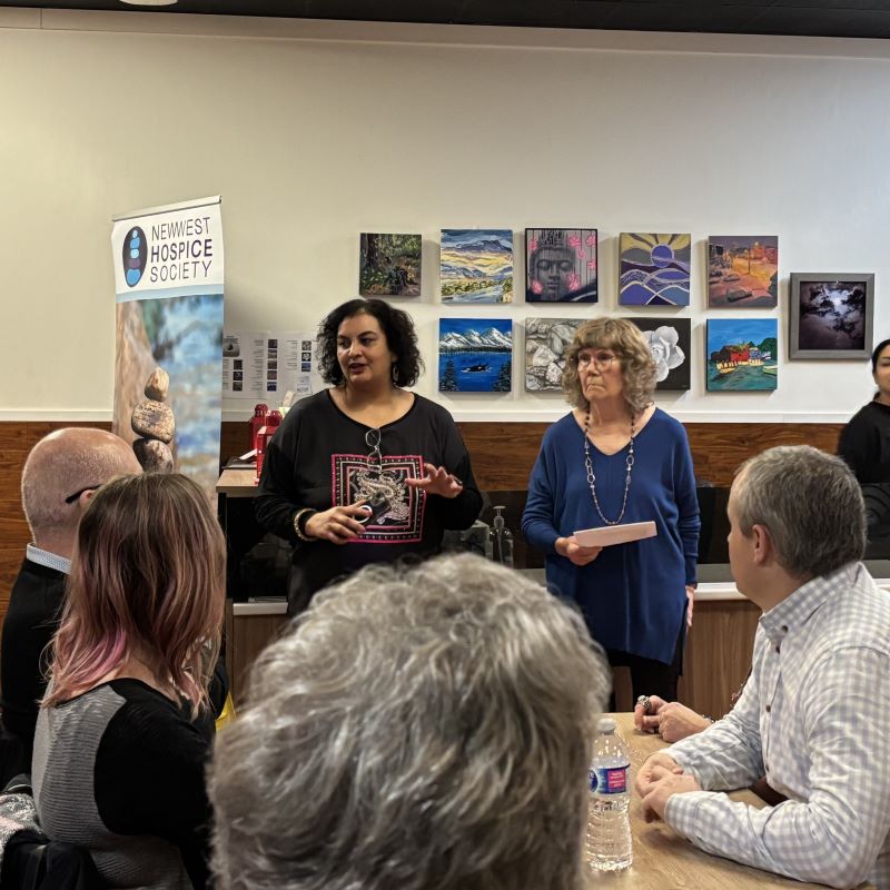 Ruby and Wendy stand in a restaurant and address the assembled diners in front of a New West Hospice banner. 